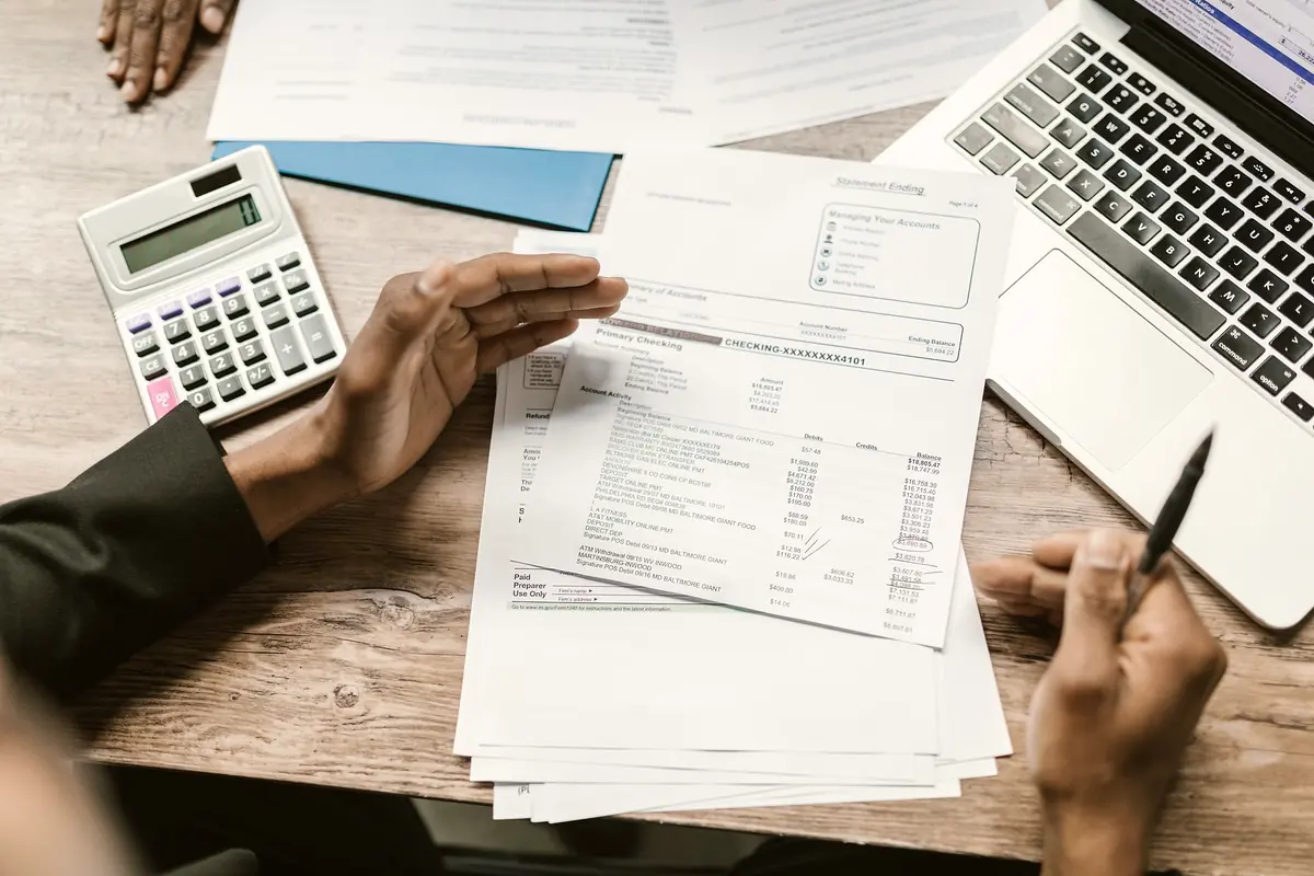 Hands checking printed account statements against a calculator and a laptop on a wooden desk.