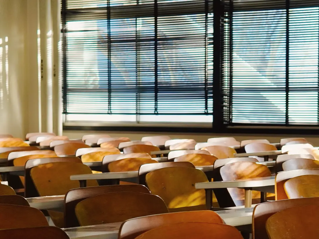 Rows of empty wooden lecture-hall seats and folding desks, lit by daylight through window blinds.